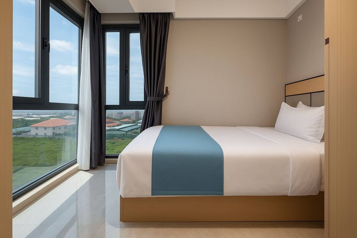 A hotel room with a large window, a bed with a blue accent, and a view of buildings and greenery outside.