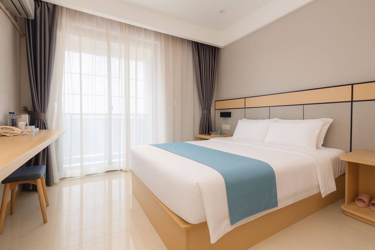 A hotel room with a bed, bedside table, desk, chair, and a window with curtains.