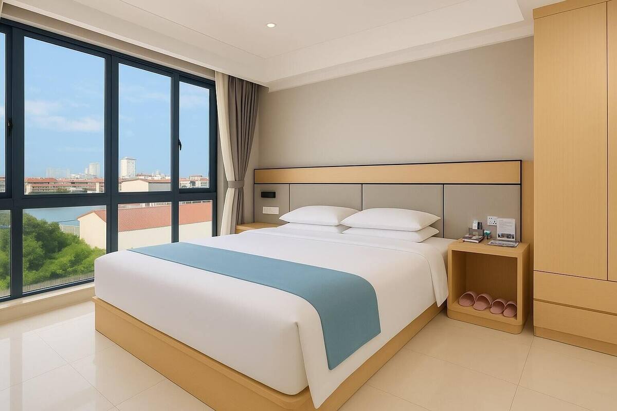 A modern hotel room with a large bed, a nightstand, a wardrobe, and a view of the city through large windows.
