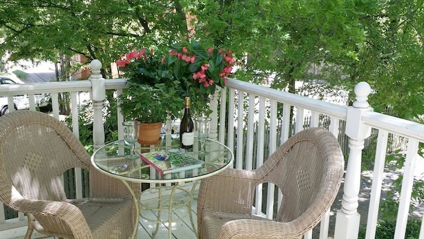 A glass-top table with wicker chairs on a balcony, surrounded by lush greenery and a white railing.