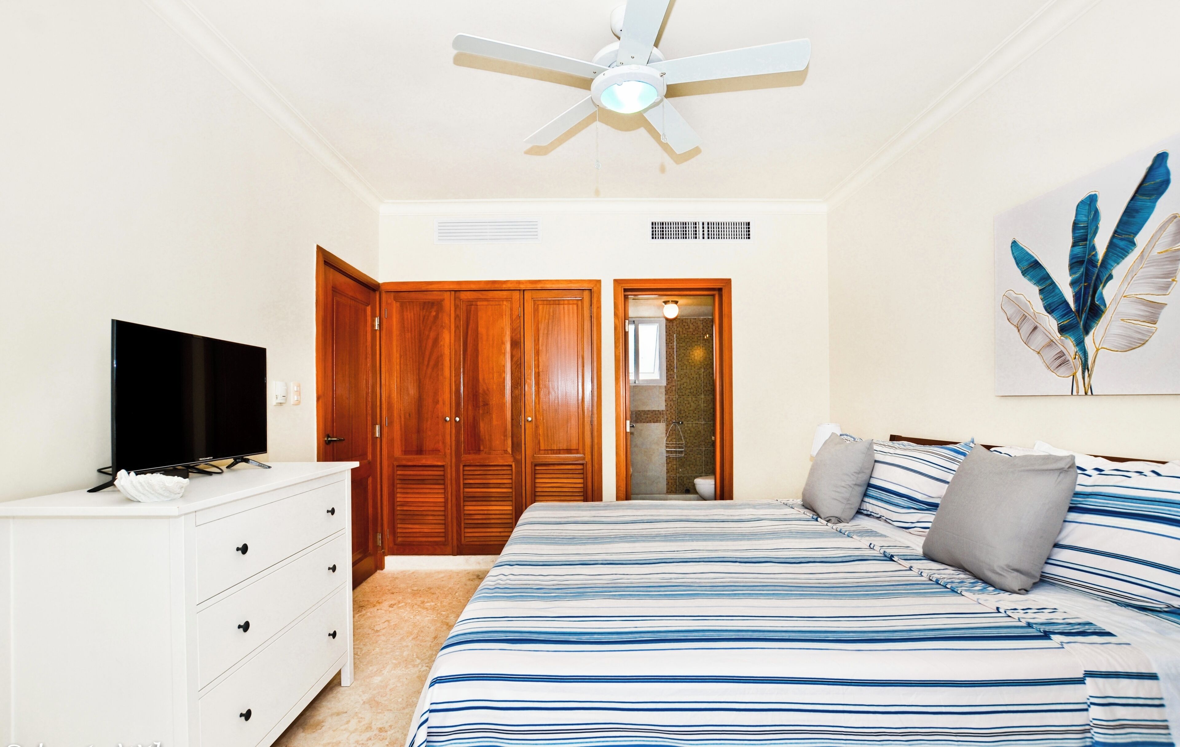 Ein Schlafzimmer mit einem Bett, einem Fernseher auf einer Kommode, Holzschränken, einem Deckenventilator und einer Duschkabine.