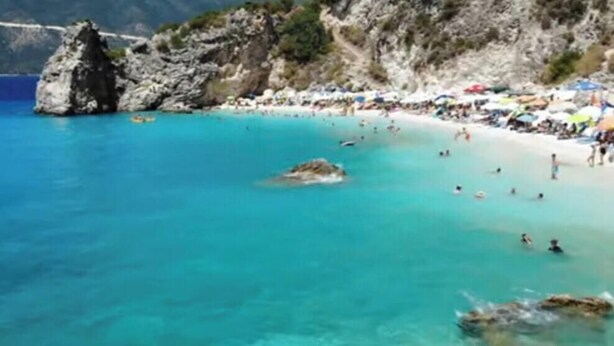 A beach with clear turquoise water, rocky formations, and a crowd of people.