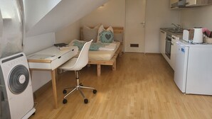 A compact, well-lit room with a wooden desk, a white chair, a bed with a wooden headboard, a kitchenette with a stove and a small refrigerator, and a ceiling fan. - Stunning 1-Bed Apartment in Bodolz Lake Constance (Bodolz)