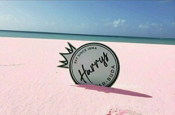 A beach with a sign on the sand that reads "Harry's Bar-Buda."