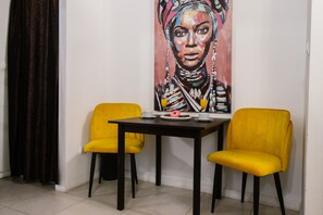 A dining area with a table and two yellow chairs, a painting on the wall, and a dark curtain.