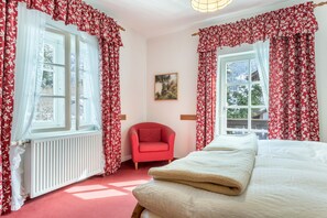 Ett sovrum med en säng, en röd fåtölj och två fönster med blommiga gardiner.