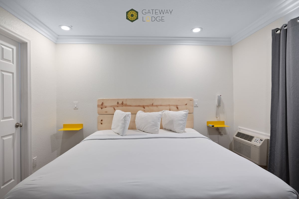 Ein Hotelzimmer mit einem hölzernen Kopfteil, weißer Bettwäsche, einem Nachttisch mit Lampe und einem an der Wand befestigten Telefon.