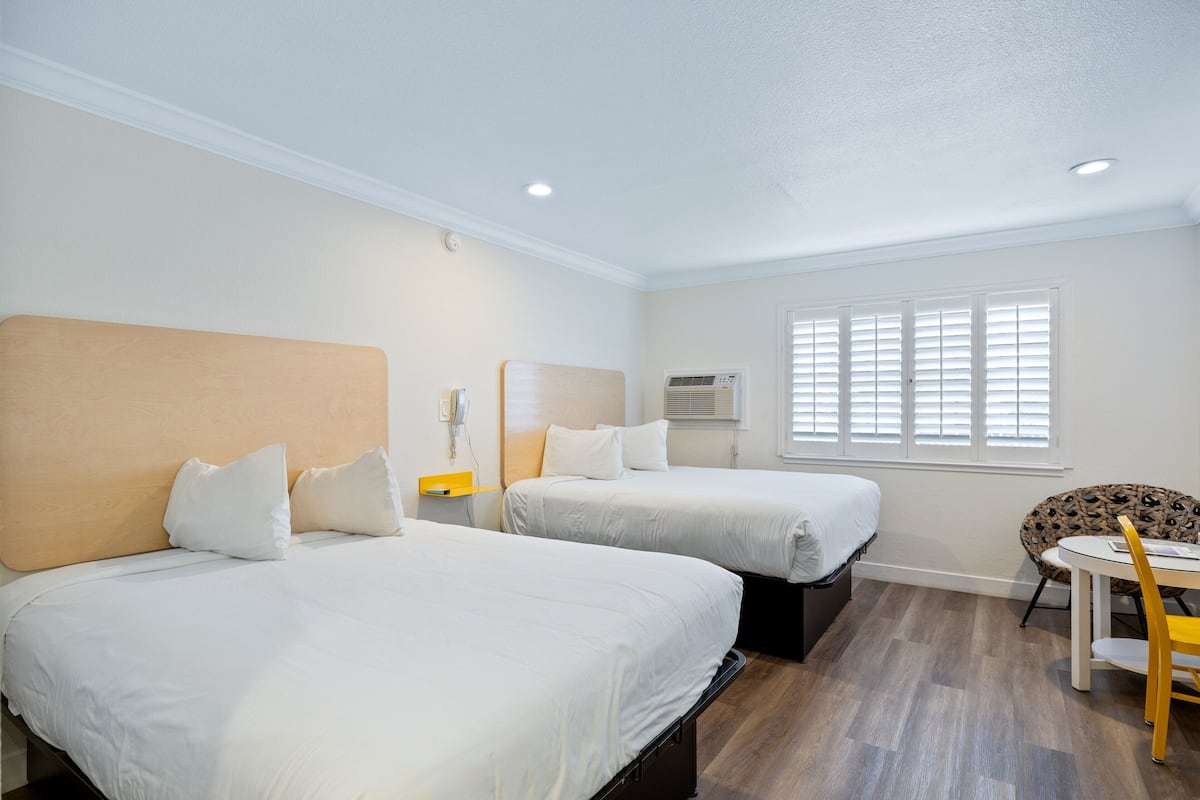 Ein Hotelzimmer mit zwei Betten, einem kleinen Tisch, einem Stuhl und einem Fenster mit Jalousien.