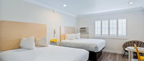 A hotel room with two beds, a small table, a chair, and a window with blinds.
