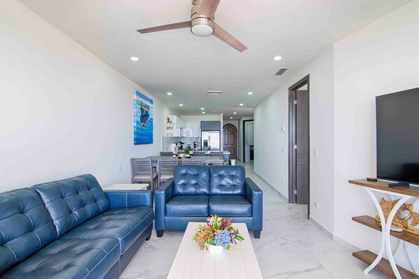 A modern living room with a blue leather sofa, a coffee table with flowers, a ceiling fan, and a TV on a wooden stand. - Casa Blanca Golf Villas 2 Bedrooms Condo E 707 (Puerto Penasco)