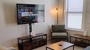 A living room with a flat-screen TV displaying a schedule, a sofa, a coffee table, and a window with blinds. - The Argyle on Mass Ave (Indianapolis)