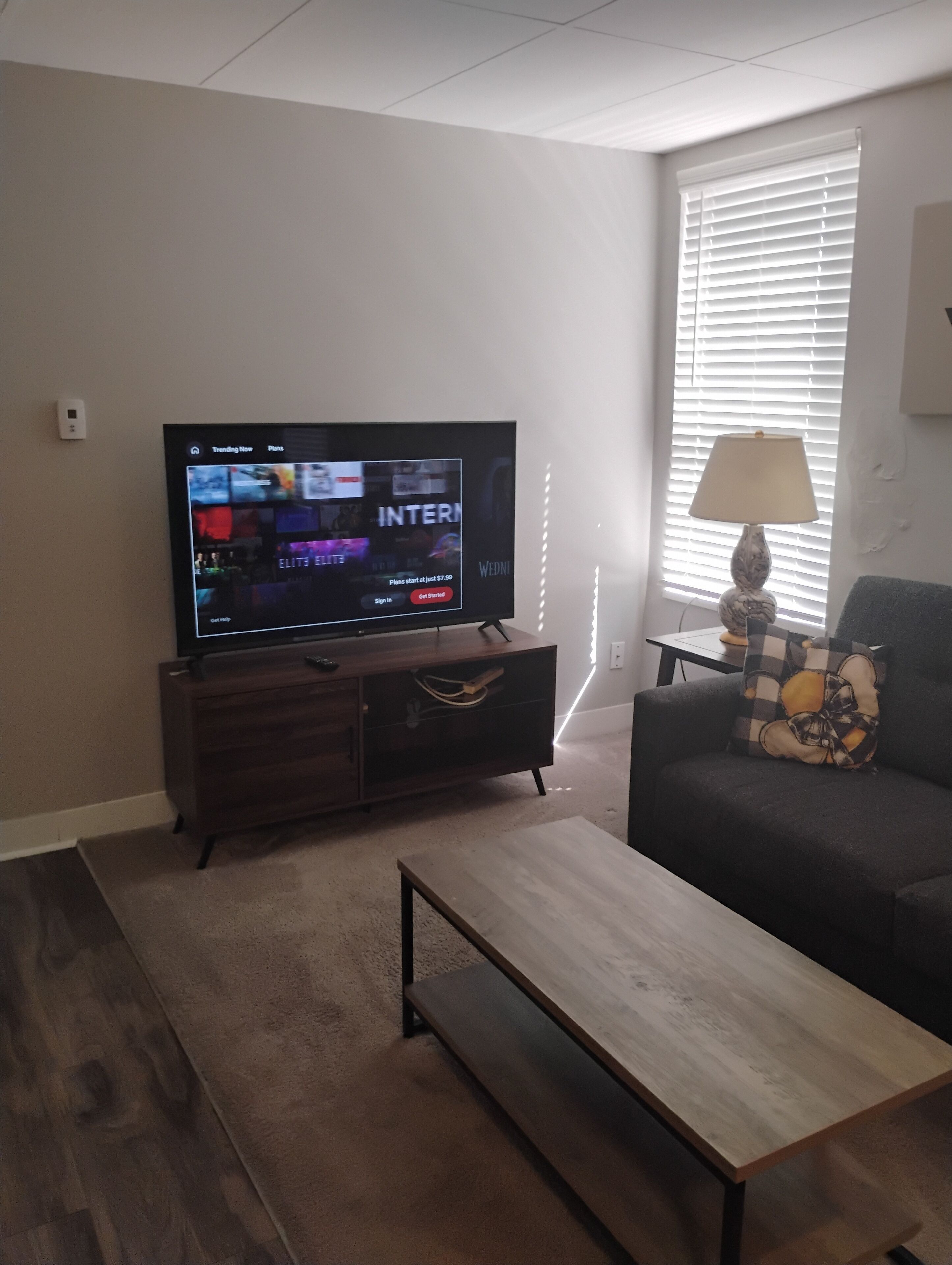 Un salon avec un téléviseur à écran plat posé sur un meuble en bois, un canapé foncé agrémenté d’un coussin à motifs, une table basse en bois et une fenêtre avec des stores.