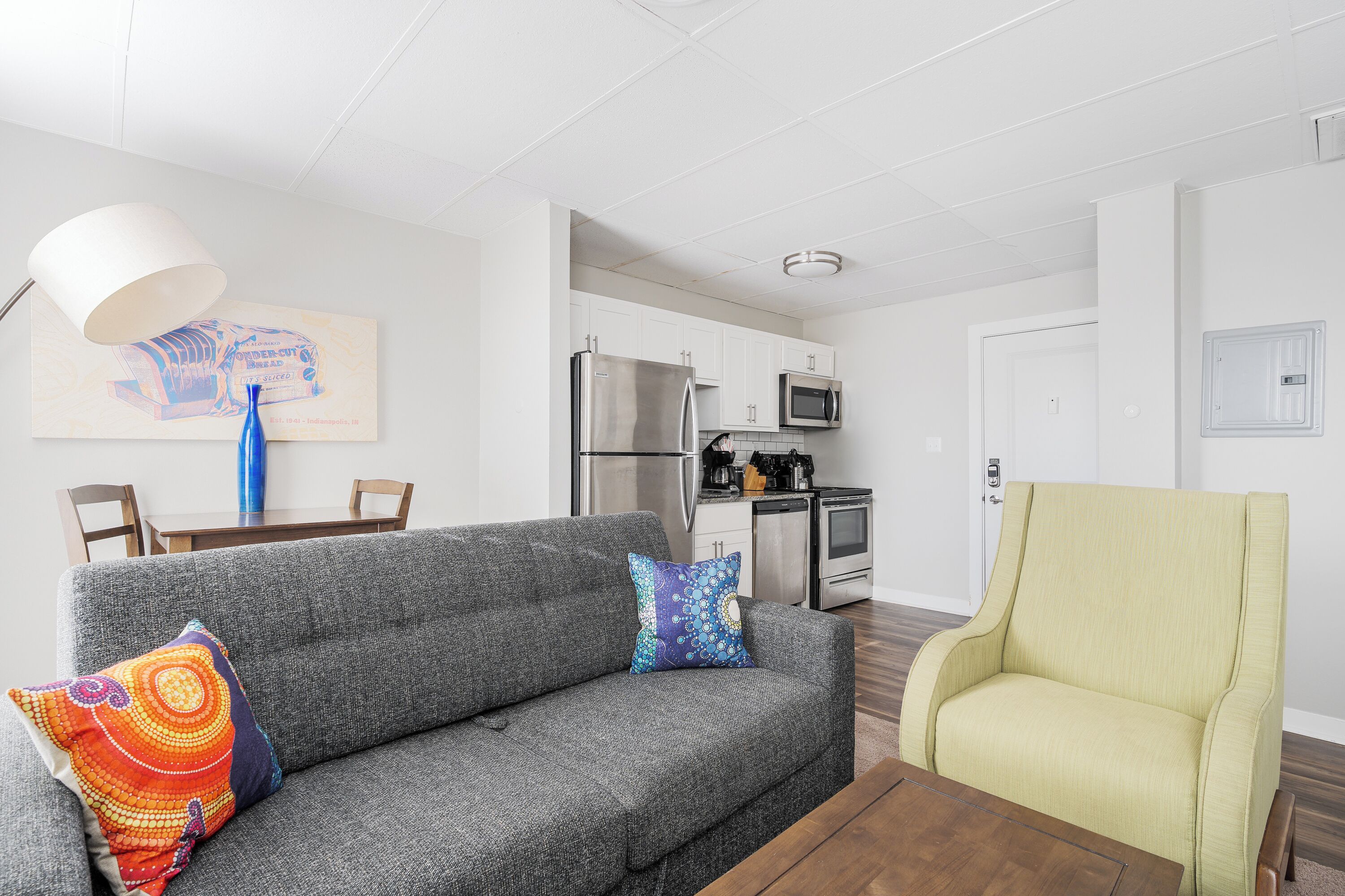 A living room with a grey sofa, a yellow armchair, and a dining table with chairs. There is a kitchen area with a refrigerator, microwave, and stove.