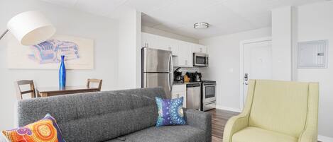 A living room with a grey sofa, a yellow armchair, and a dining table with chairs. There is a kitchen area with a refrigerator, microwave, and stove.