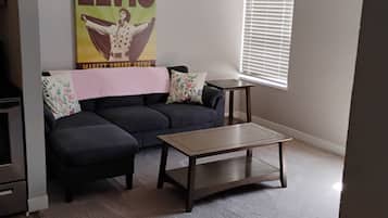 A living room with a dark sofa, a coffee table, a wall poster, a window with blinds, and a ceiling fan.