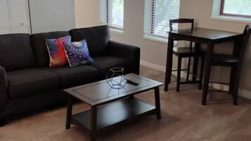 Un salon avec un canapé marron foncé, une table basse assortie et une table à manger en bois avec des chaises. Il y a des fenêtres avec des stores, une porte et un sol recouvert d’une moquette.