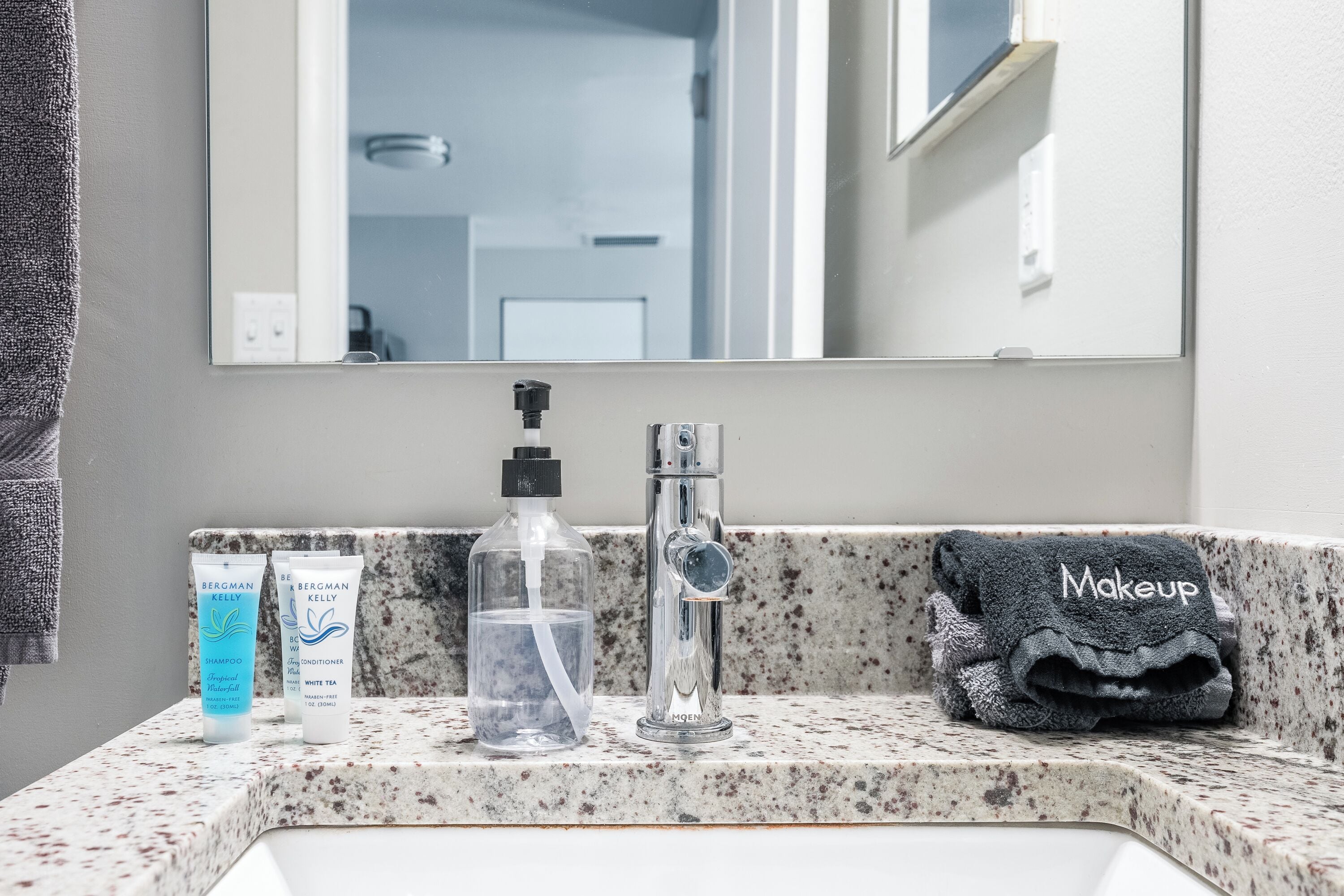 Un lavabo de salle de bain avec un miroir, un flacon de shampoing, un flacon d’après-shampoing, un flacon de thé blanc et une serviette sur laquelle est écrit le mot « Maquillage ».