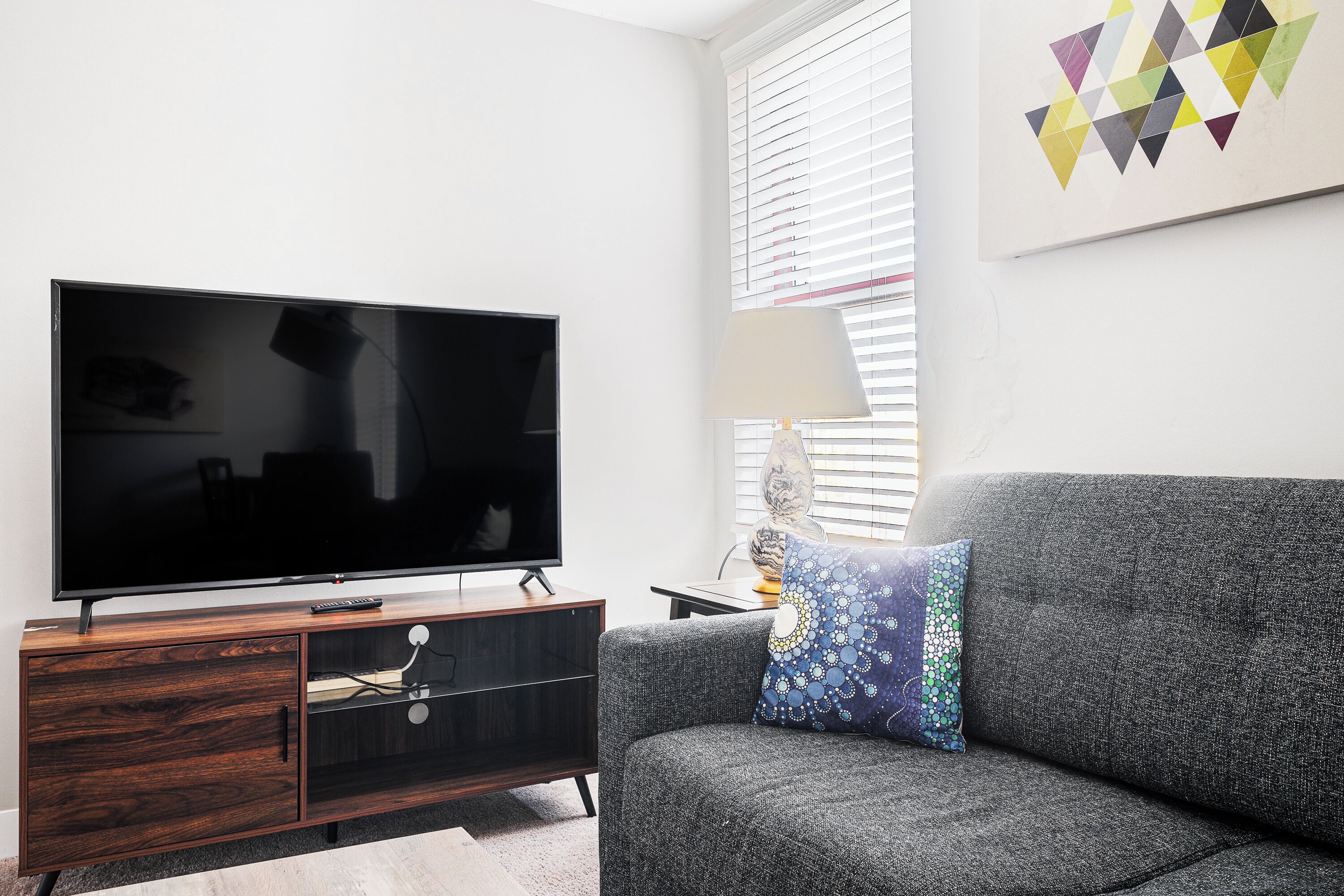 A modern living room with a flat-screen TV on a wooden cabinet, a grey sofa, and a patterned pillow.