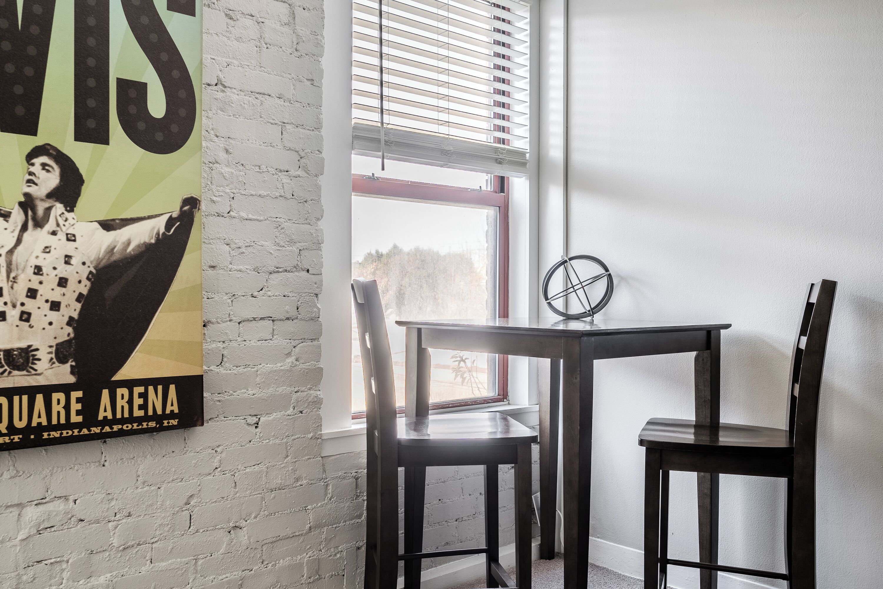 A room with a white brick wall, a poster of a performer, a window with blinds, a round table, and two chairs.