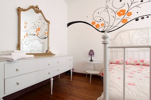 Una camera da letto con una cassettiera bianca, un letto con copriletto floreale, uno specchio decorativo e un tavolino con una lampada viola.
