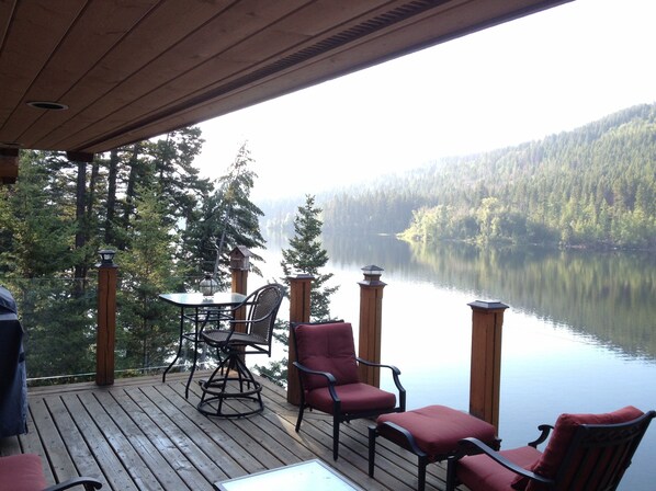 A wooden deck with a table and chairs overlooking a calm lake and forested hills.