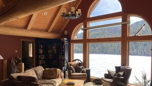 A cozy cabin interior with a large window overlooking a lake, a fireplace, and a collection of bookshelves.