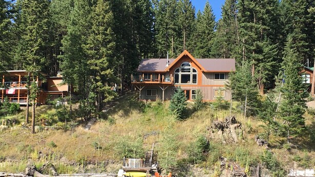 A large wooden cabin with a balcony, surrounded by dense forest and a dock extending into a lake.