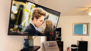 Una sala de estar moderna con un televisor de pantalla plana mostrando 'Twilight', un sofá gris, una mesita con un control remoto y un mueble negro.