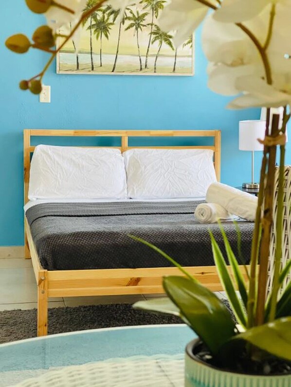 A bed with white linens, a dark gray blanket, and a rolled towel. There is a painting of palm trees on the wall behind the bed.