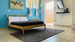 Un dormitorio con cama, un cuadro de palmeras, una ventana con cortinas, un equipo de aire acondicionado y un televisor en un estante.