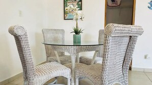 Un comedor con una mesa redonda de vidrio rodeada por cuatro sillas de mimbre blanco. Hay una planta en maceta con flores blancas sobre la mesa.