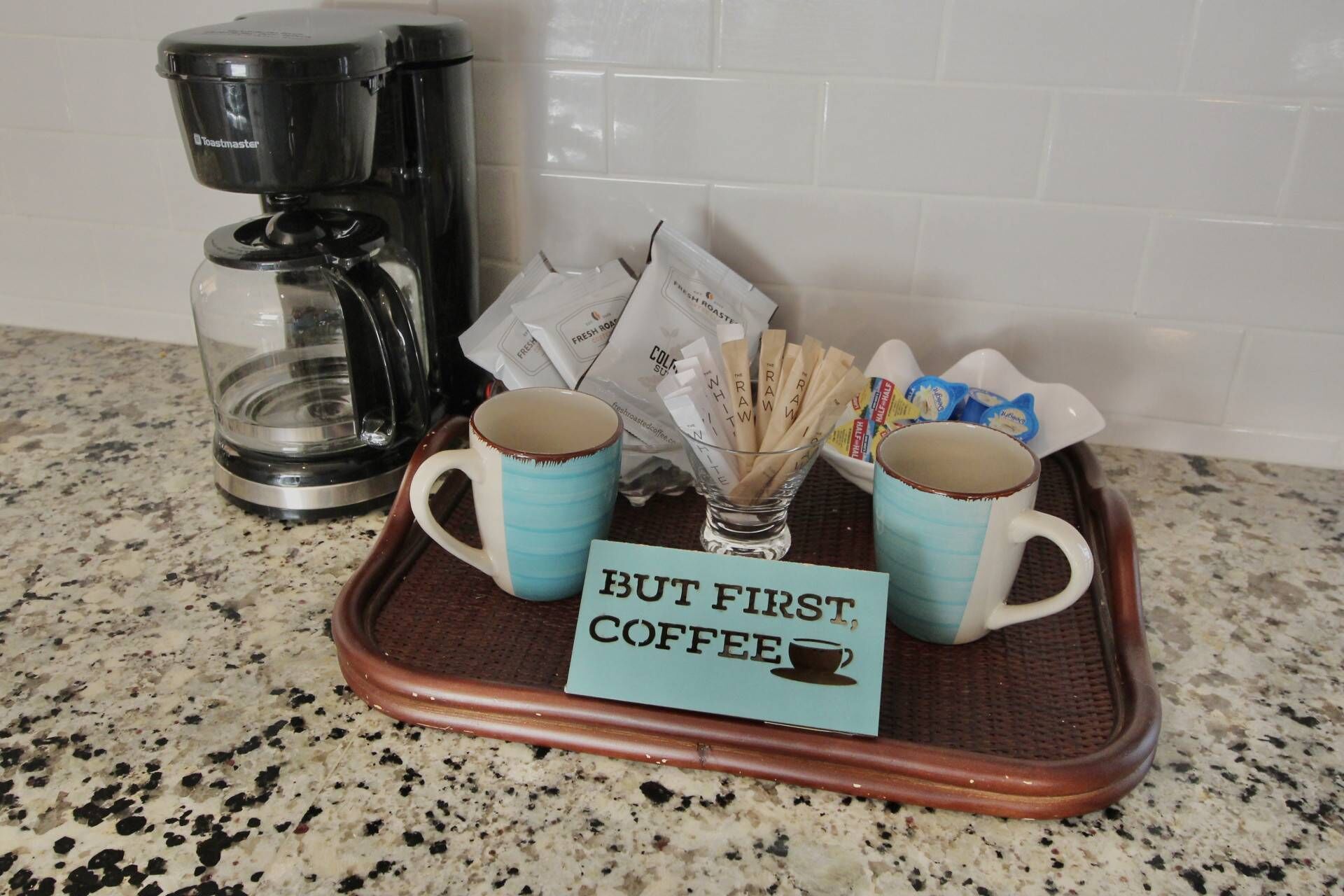 A tray with two coffee mugs, a glass of water, coffee pods, and a sign that reads "BUT FIRST COFFEE".