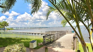 A wooden boardwalk leading to a bridge over water, with a white picket fence and palm trees.