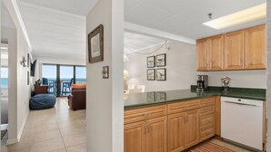 A modern kitchen with wooden cabinets, a dishwasher, and a coffee maker. There is a dining area with a table and chairs, and a view of the ocean through large windows.
