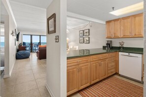 A modern kitchen with wooden cabinets, a dishwasher, and a coffee maker. There is a dining area with a table and chairs, and a view of the ocean through large windows.