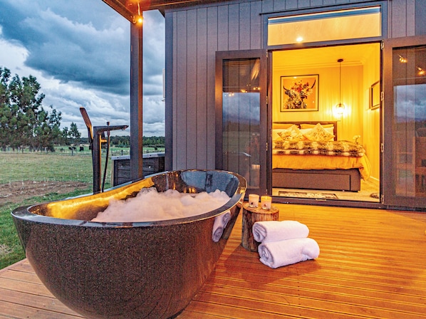 A modern outdoor hot tub with a wooden deck, a cozy bedroom with a large bed and a painting, and a glass door leading to the bedroom.
