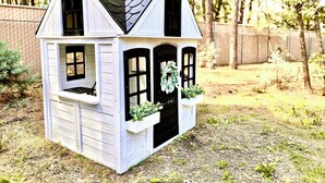 Una casita de juegos de madera blanca con techo negro, dos ventanas y una puerta adornada con un adorno de guirnalda.