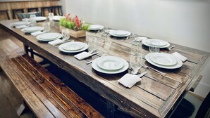 Una larga mesa de madera para comer, con platos blancos, vasos y cubiertos, flanqueada por bancos.