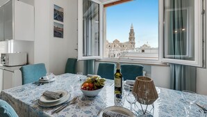 Un comedor con una mesa dispuesta para dos personas, un cuenco de fruta, una botella de vino y vistas a un edificio histórico a través del ventanal.