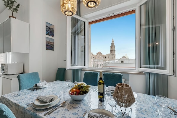 A dining area with a table set for two, a bowl of fruit, a bottle of wine, and a view of a historic building through the window.