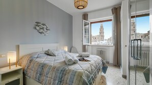 Un dormitorio moderno con una cama grande, una mesita de noche con una lámpara, una ventana con vista a la ciudad y una puerta de vidrio que da a un balcón.