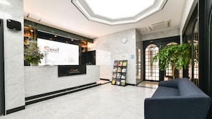 Ein Hotel-Empfangsbereich mit einer Rezeption, einer digitalen Infotafel, einer Sitzecke und einer Pflanze.