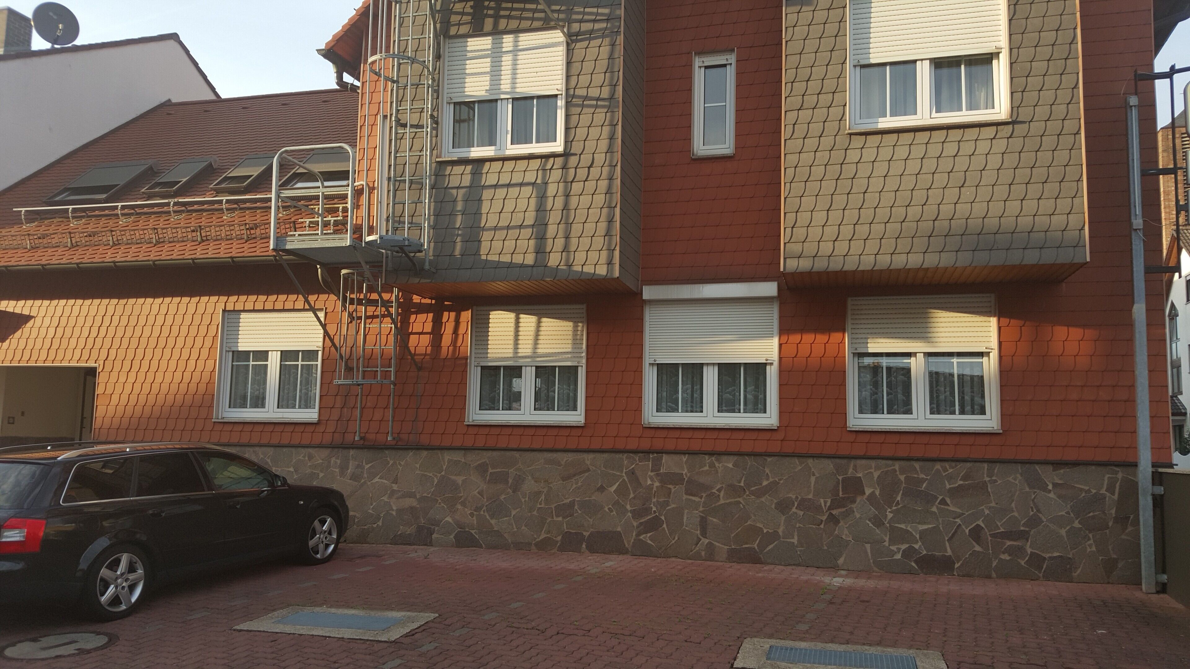 A multi-story building with a stone facade, multiple windows with shutters, and a car parked in front.