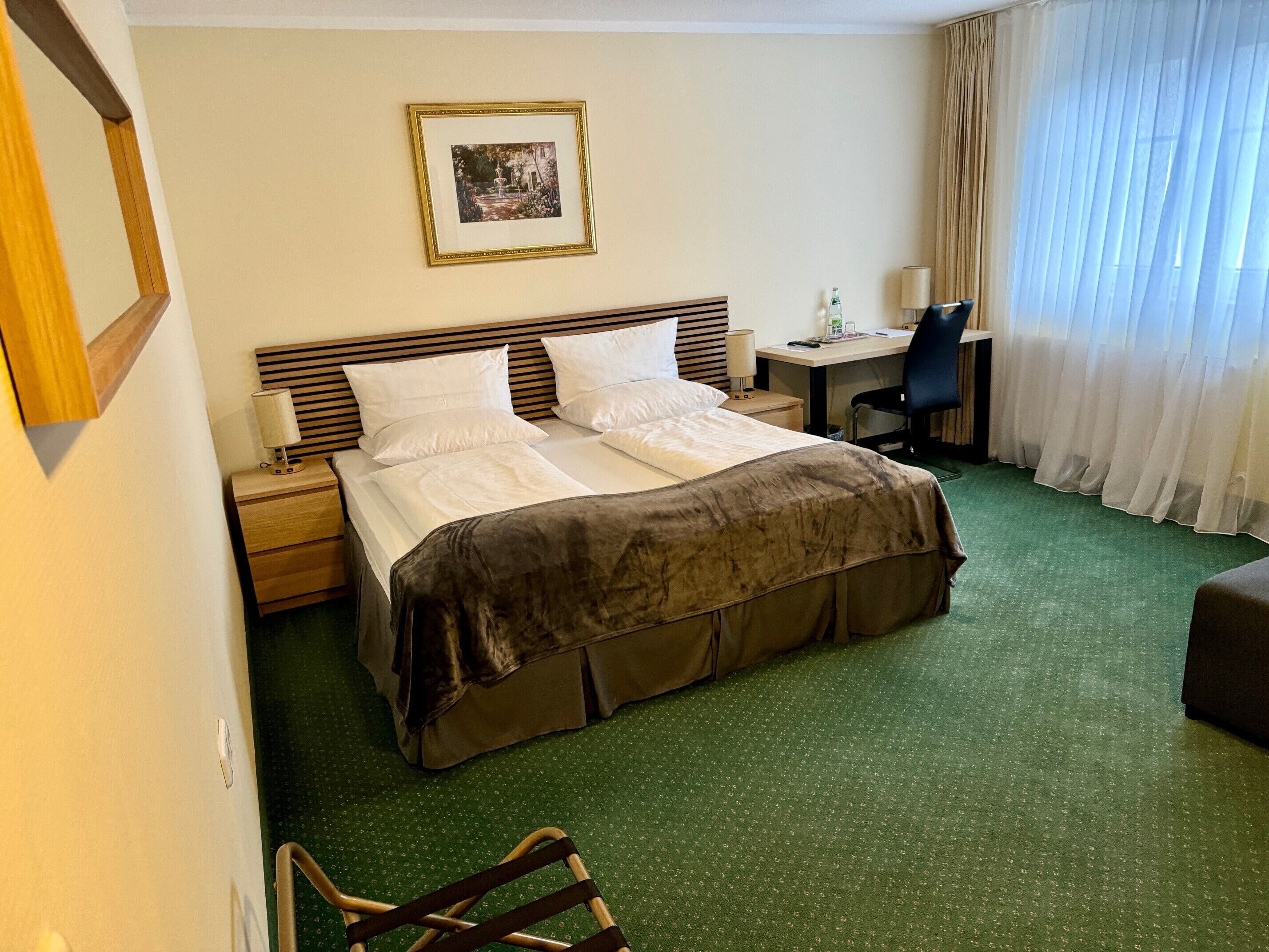 A hotel room with a bed, a desk, a chair, a picture on the wall, and a window with curtains.