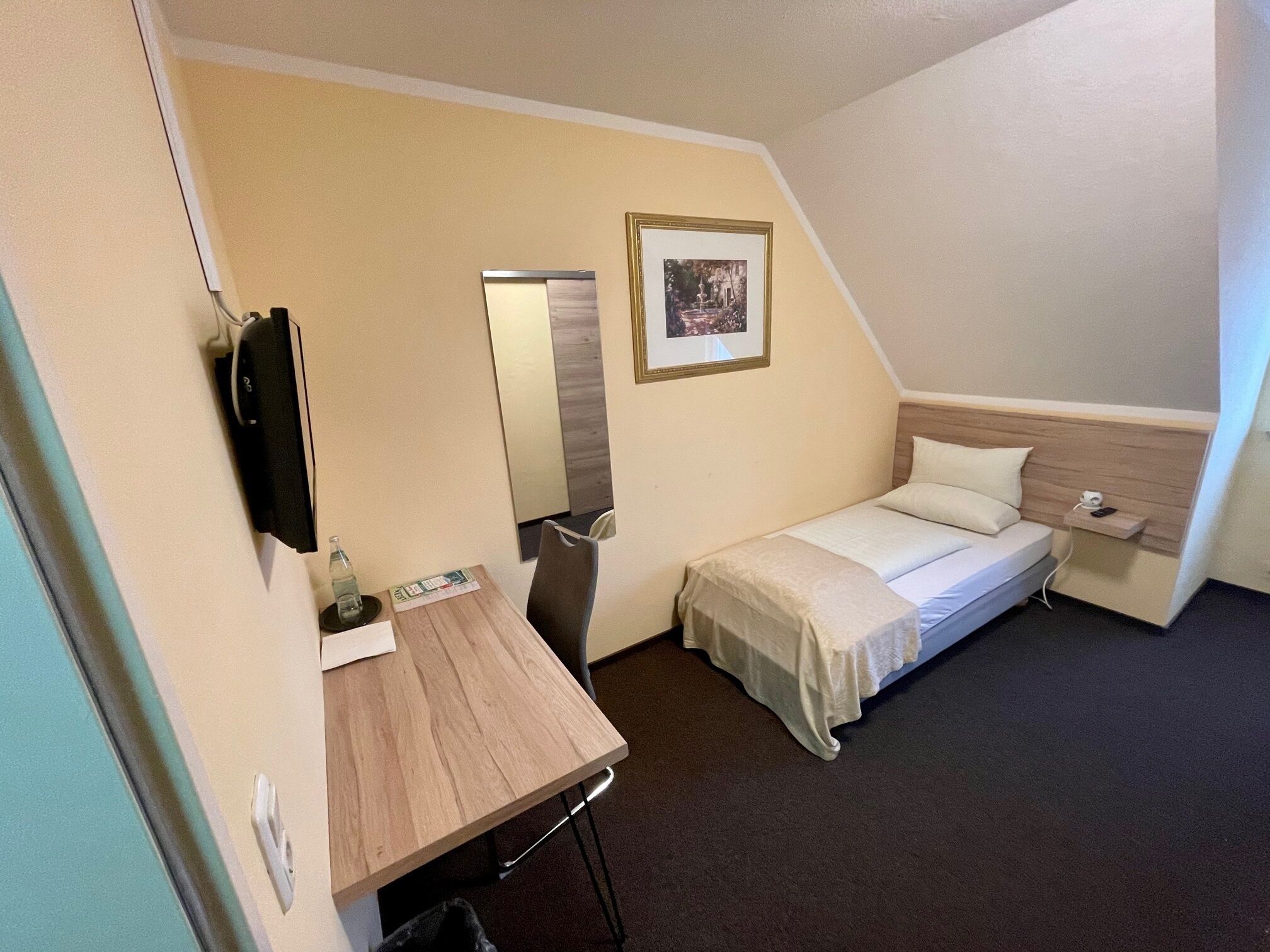 A small, compact hotel room with a single bed, a desk with a chair, a television, and a framed picture on the wall.
