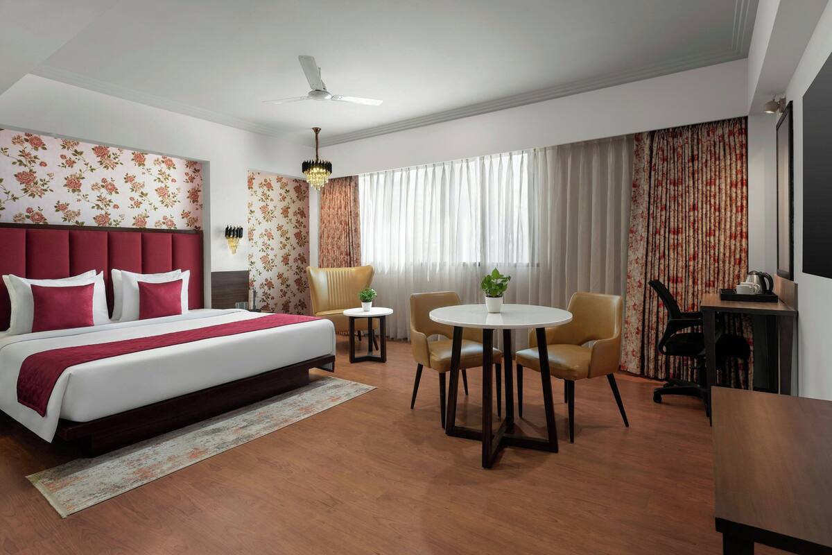 A hotel room with a large bed, a round dining table, two chairs, a small side table, a ceiling fan, and a large window with curtains.