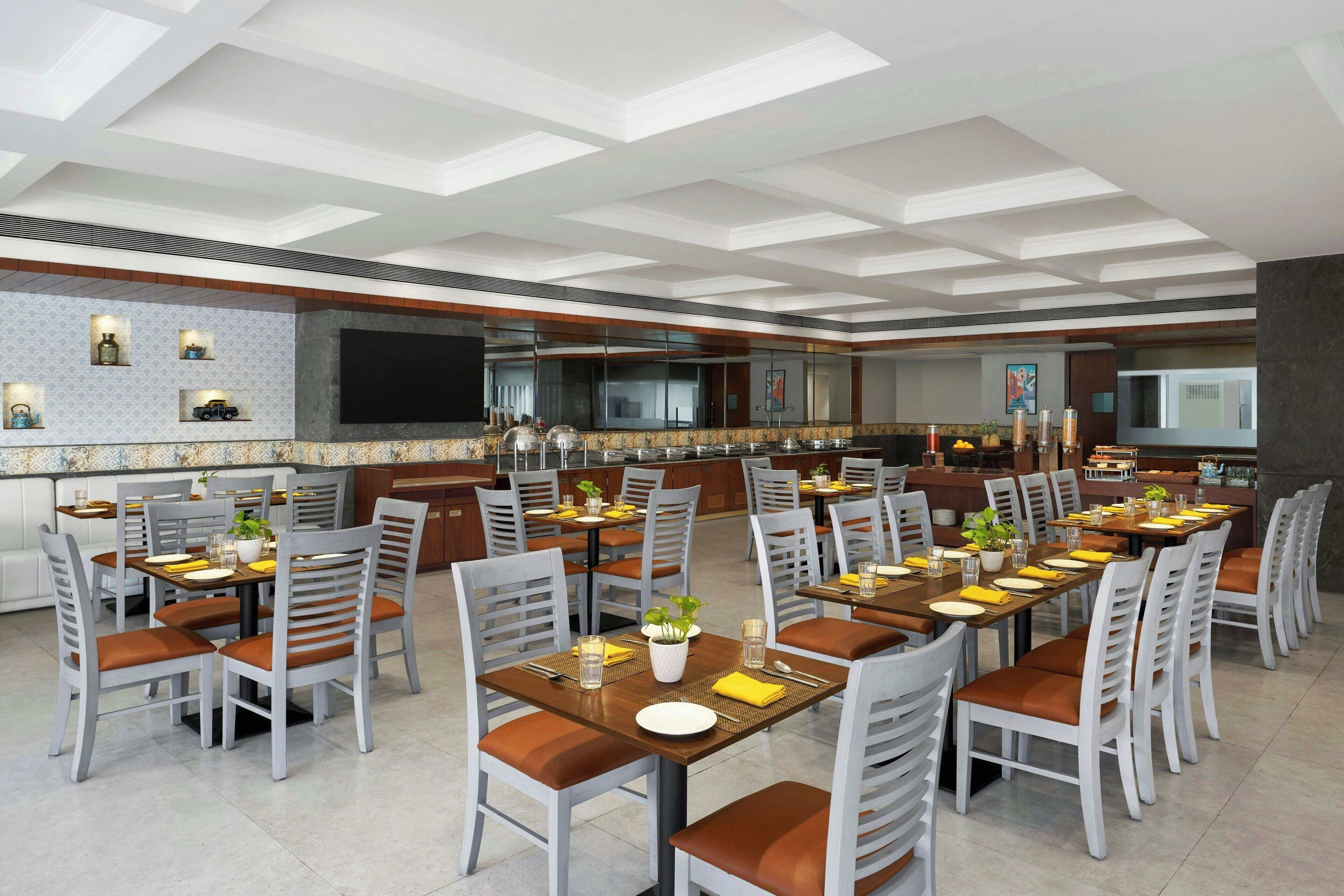 A spacious dining area with multiple tables and chairs, a kitchen in the background, and a modern ceiling design.