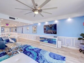 Een ruime woonkamer met een plafondventilator, een grote televisie en een blauw, patroonrijk vloerkleed.