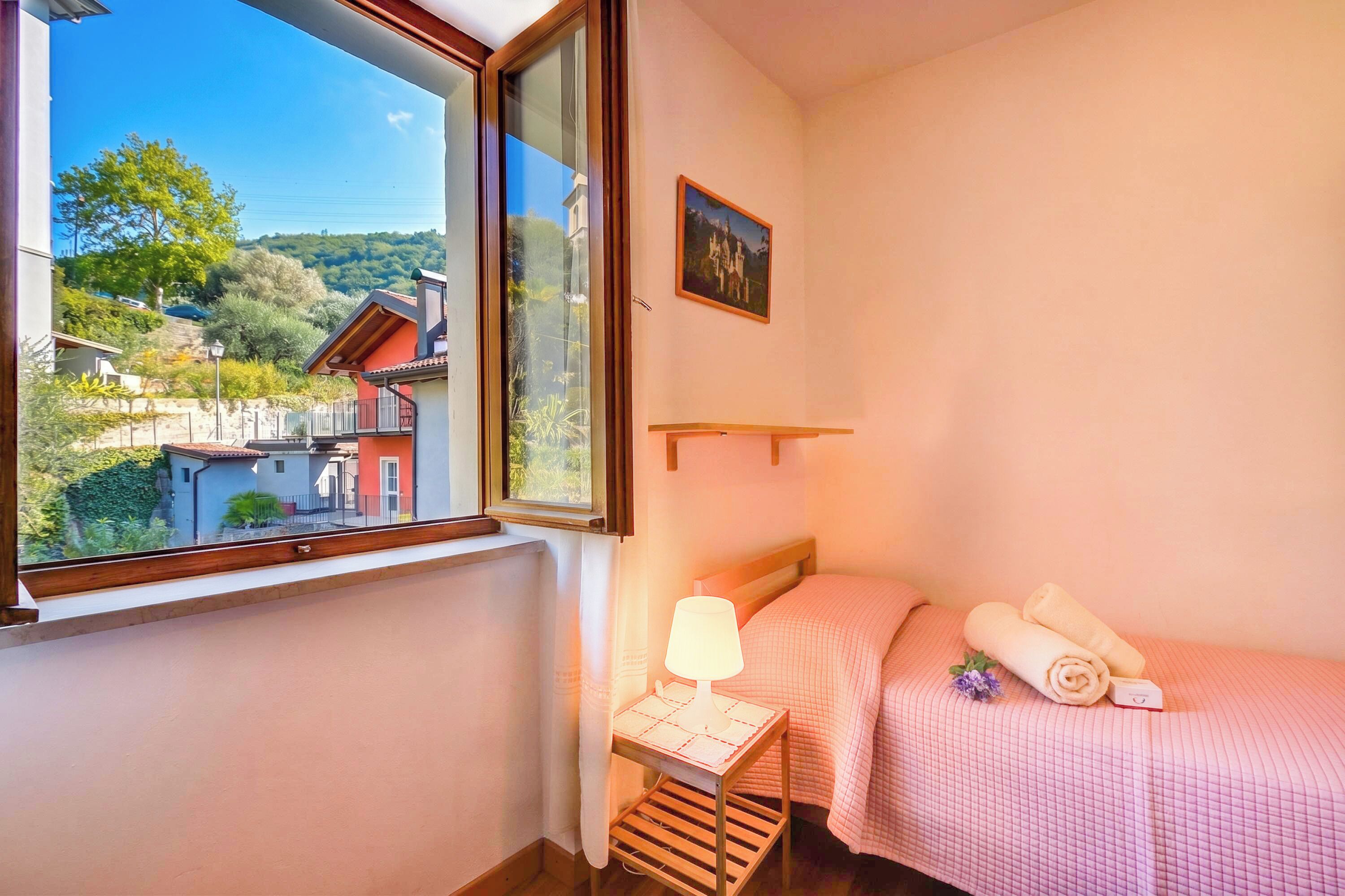 Een slaapkamer met een raam, een nachtkastje met een lamp, een bed met roze beddengoed en een lijst met een afbeelding aan de muur.