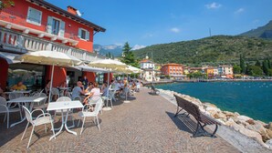 Un café frente al agua con terraza, un paseo empedrado de piedra y edificios al fondo.
